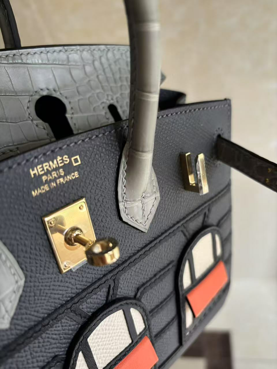 An angled close-up view of the Hermes Faubourg Birkin 20 Rainy Day, showing the 'HERMÈS PARIS MADE IN FRANCE' gold heat stamp, the turn-lock, and the textured Gris Misty Epsom leather.