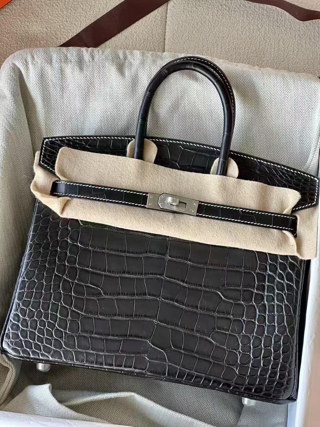 A black alligator Birkin with contrast white stitching and silver hardware, resting in its original dustbag and packaging.