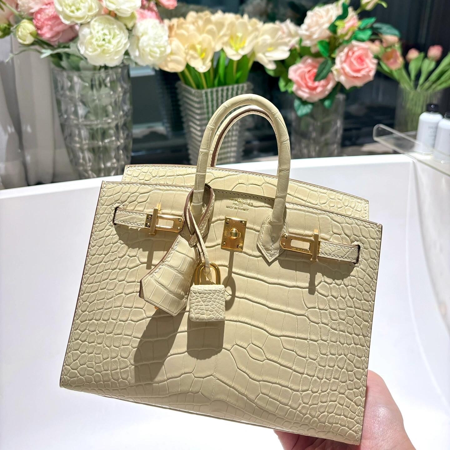 An open cream-colored alligator Birkin bag with gold hardware, lock, and clochette, displayed beautifully in front of a floral background.
