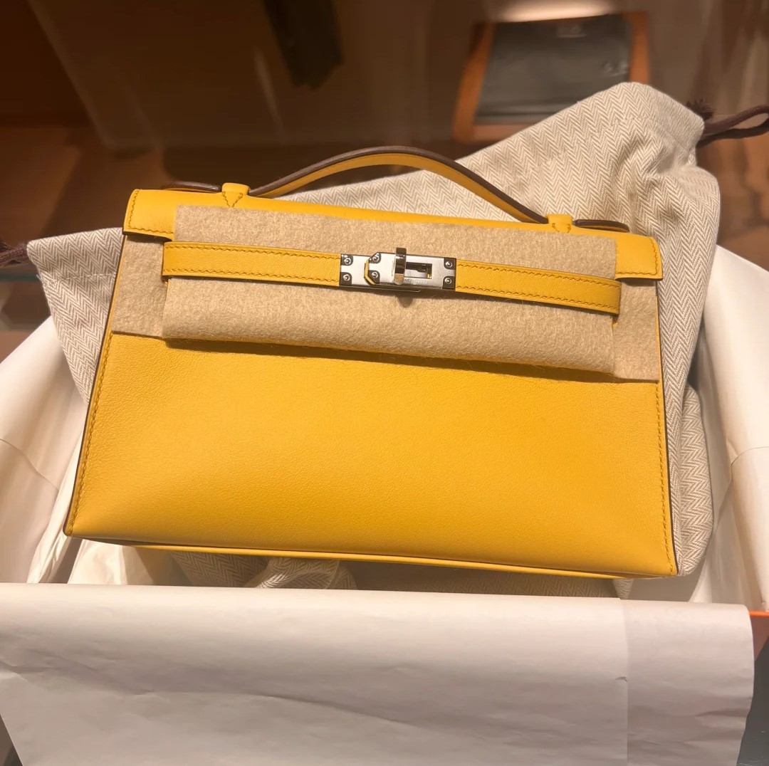 A bright yellow leather Hermès Kelly Pochette bag with palladium hardware resting on a protective felt cloth inside the box.