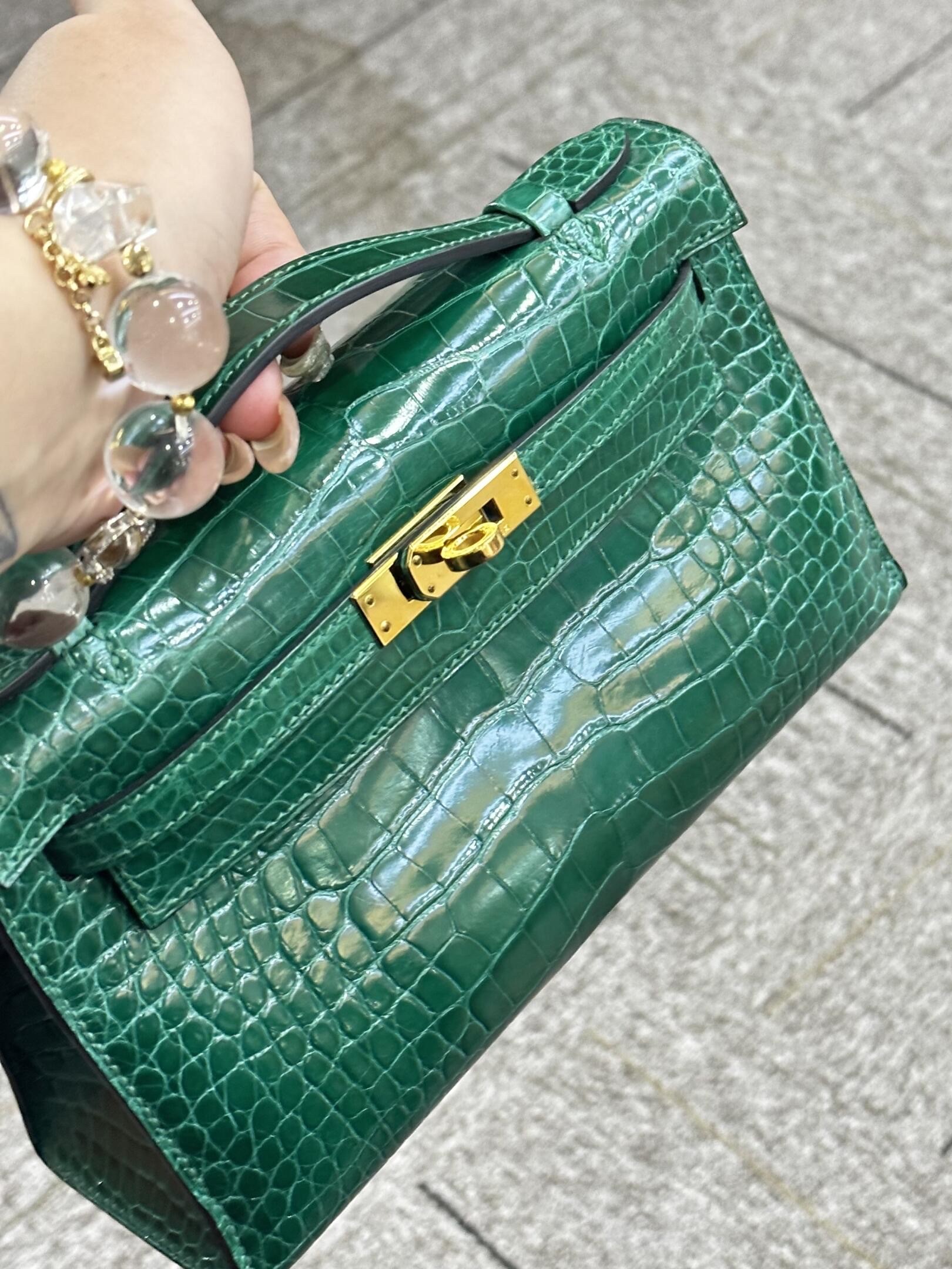 Close-up buyer show of a luxury Hermès Kelly Pochette clutch in shiny green alligator exotic leather with gold hardware.