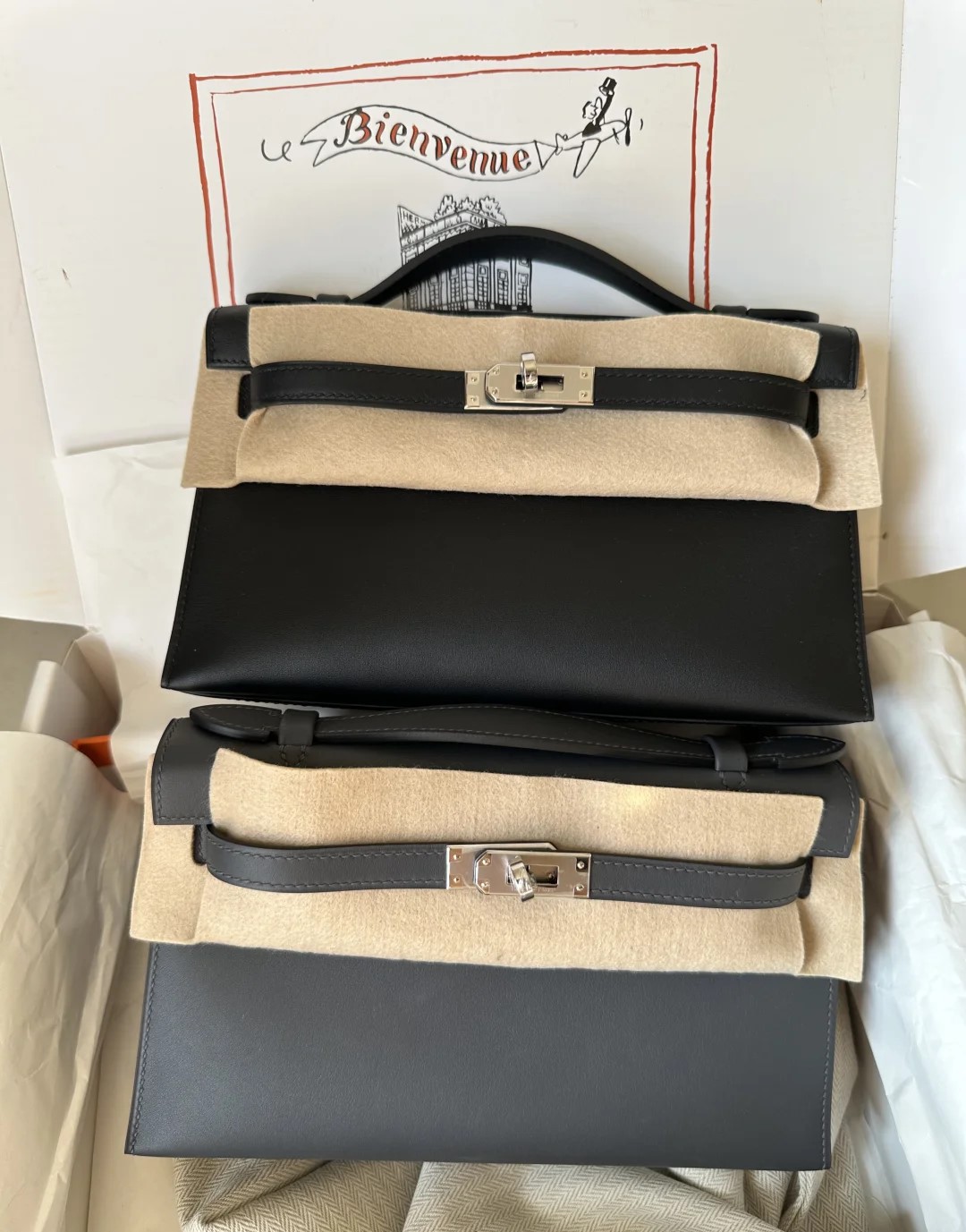 Two Hermès Kelly Pochette clutch bags, one in black and one in dark grey leather, showcased together in an original box with protective felt.