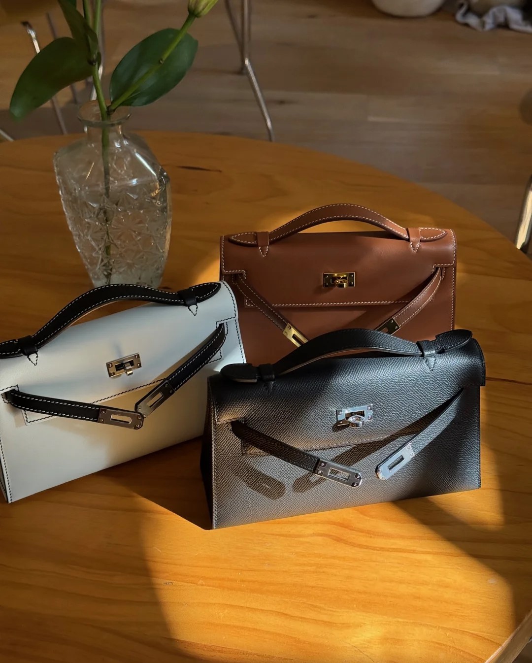 A collection of three Hermès Kelly Pochette bags in white with black stitching, classic brown, and textured black leather on a wooden table.