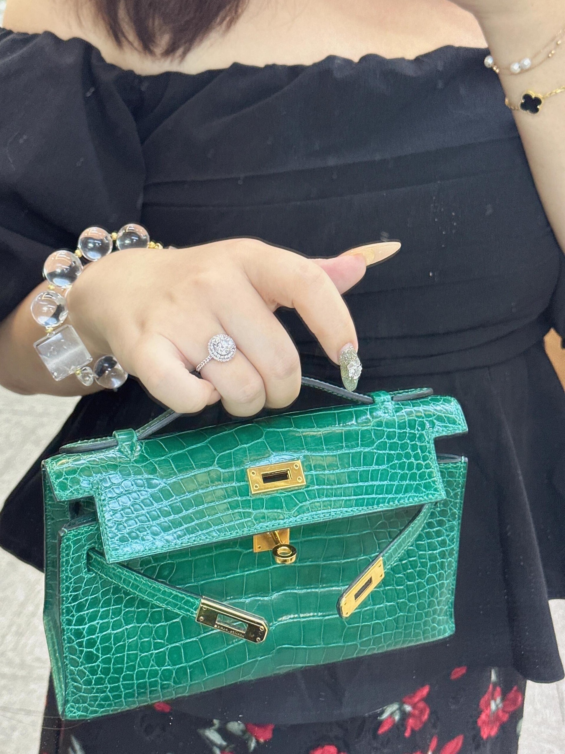 A buyer wearing a black dress holding a green exotic leather Hermès Kelly Pochette bag with gold-tone hardware.