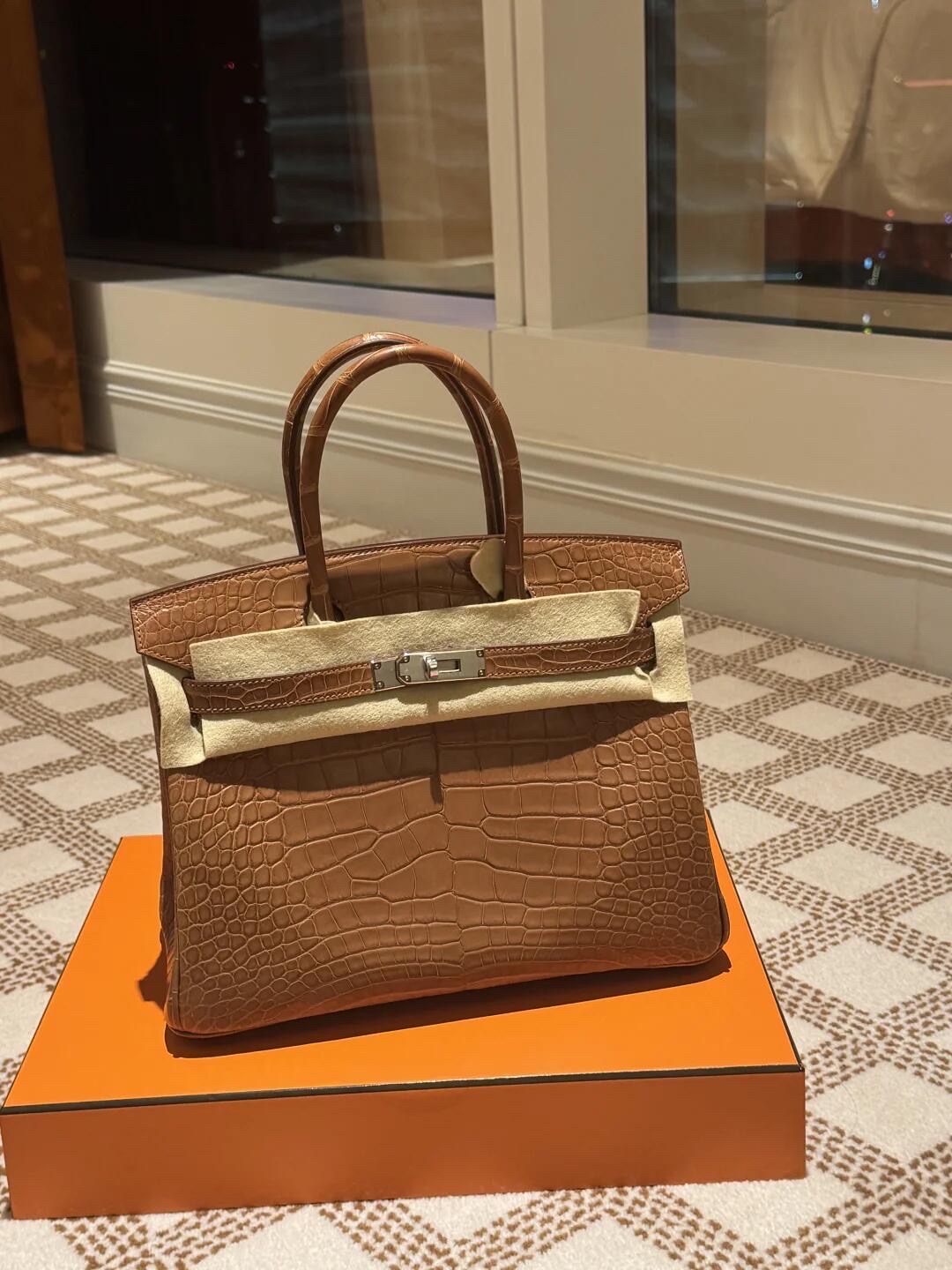 A classic tan brown Birkin alligator bag with silver hardware and a protective felt layer, sitting on top of an iconic orange Hermès box.